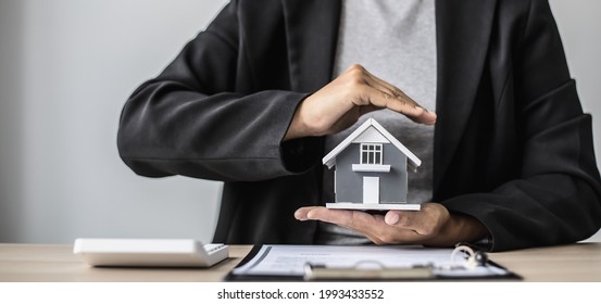 A Small Gray Model House Is An Example Of A Home In A Housing Project, An Insurance Salesperson Is Doing A Symbolic Home Protection Act In Home Insurance. Real Estate Leasing Concept.