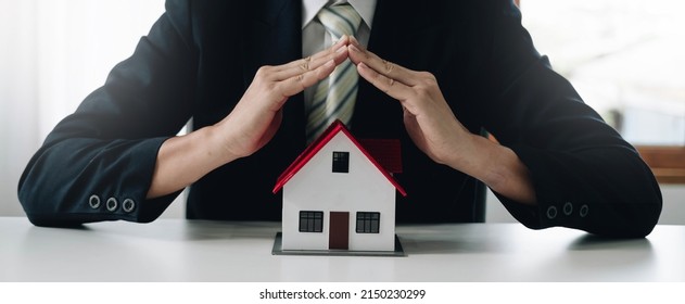 A Small Gray Model House Is An Example Of A Home In A Housing Project, An Insurance Salesperson Is Doing A Symbolic Home Protection Act In Home Insurance. Real Estate Leasing Concept.
