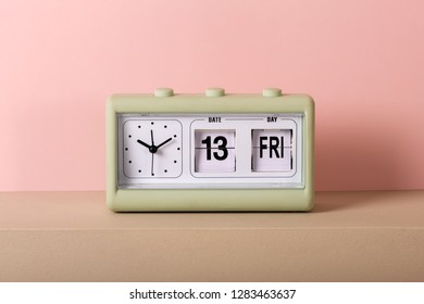 Small Green Vintage Clock With White Face And Calendar Showing Friday 13th. Viewed From The Front In Close-up, Against Pale Pink Background