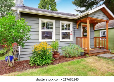 Small Grey House With Wooden Deck. Front Yard With Flower Bed And Lawn