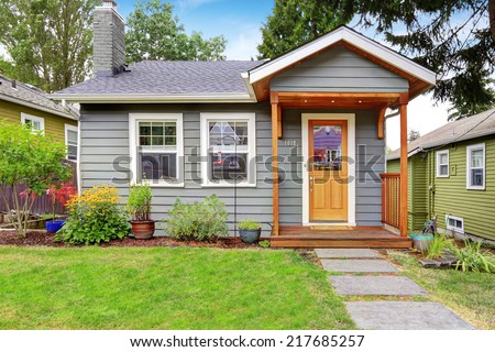 Small grey house with wooden deck. Front yard with flower bed and lawn