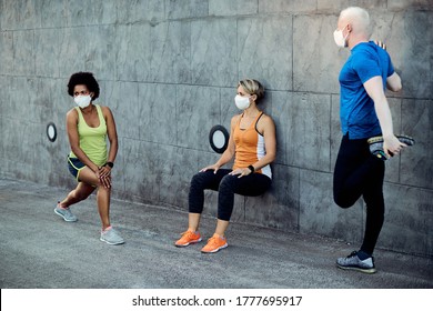 Small Group Of Athletic People Wearing Protective Face Masks And Doing Relaxation Exercises While Warming Up Before Running Outdoors. 