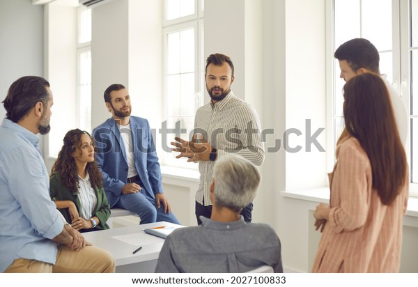 Small group of diverse women and men listen to a\
confident male corporate business coach. Man advises clients,\
trains employees or explains the strategy of a marketing seminar.\
Staff training concept.