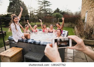 Small Group Of Female Friends Relaxing And Celebrating In A Hot Tub. They Are All Looking Towards Their Friend Who Is Taking A Group Photo Of Them.