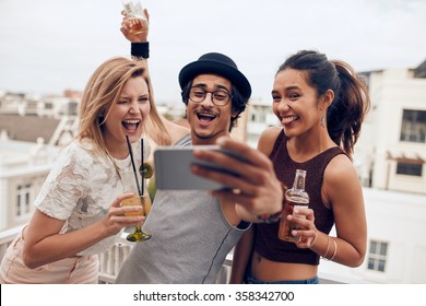 Small Group Of Friends Taking Selfie On A Mobile Phone. Young Man And Women With Drinks Making Funny Face While Taking A Self Portrait On Smart Phone. Having Fun On Rooftop Party.