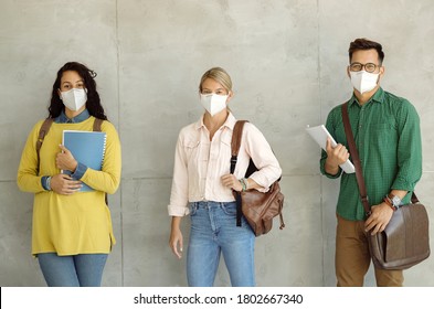 Small Group Of Happy University Students Wearing Protective Face Masks While Standing By The Wall In A Hallway And Looking At The Camera. 