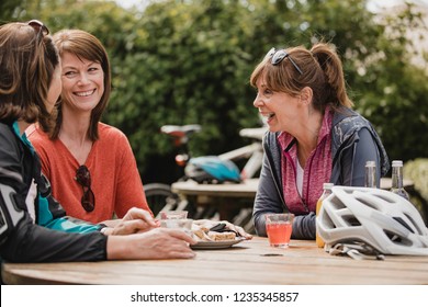 Small Group Of Mature Female Cyclists Sitting Down In The Outside Seating Area Of A Cafe. Tey Are Enjoying Some Refreshments As They Take A Break.