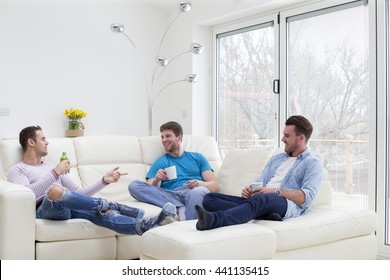 A Small Group Of Men Are Sat In A Living Room Relaxing. They Are All Talking And Smiling Drinking Beverages.