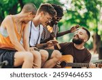 Small group of multicultural street performers sitting in city park, playing a guitar and singing.Interracial musicians sitting in public park and having jam session or rehearsal. Entertainers in park