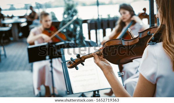 Small Group Of Musicians, Two Violinists And One
Cellist, Young Girls With Summer Lilac Dresses Playing On The
Summer Terrace