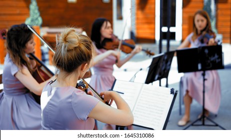 A Small Group Of Musicians, Two Violins And Cello, Beautiful Girls In A Classic Style Of Clothes Play On The Summer Terrace From The Outside