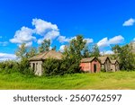 A small group of old houses sit in a grassy field. The houses are old and run down, with some of them having a brick exterior. The sky is clear and blue