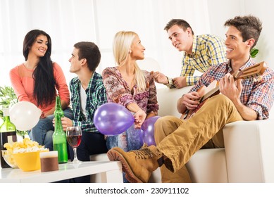 A Small Group Of Young People Hang Out At The House Party, Chatting With Each Other While Their Friend Having Fun Playing Acoustic Guitar.