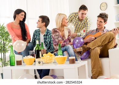 A Small Group Of Young People Hang Out At The House Party, Chatting With Each Other While Their Friend Having Fun Playing Acoustic Guitar.