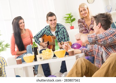 A Small Group Of Young People Hang Out At The House Party, Chatting With Each Other While Their Friend Having Fun Playing Acoustic Guitar.