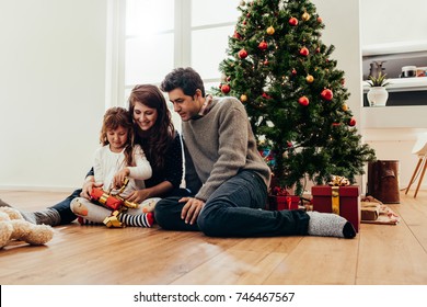 Small And Happy Family Unboxing Gifts Sitting Beside A Christmas Tree. Young Couple Watch Their Daughter Open Her Christmas Gift.