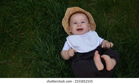 Small Happy Newborn child in summer panama hat Fall down laying on grass barefoot in Summer Sunny Day. Infant Kid Toddler Boy Smilling Face look at camera in Garden ouside Family Childhood Nature.