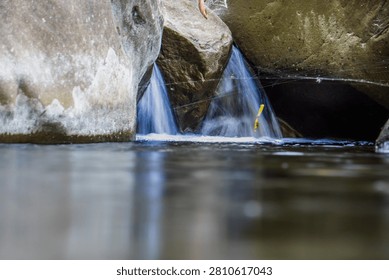 A small hidden waterfall flows smoothly through big rocks into a calm river pool in the middle of nature, creating a serene and peaceful atmosphere in a wild forest landscape.