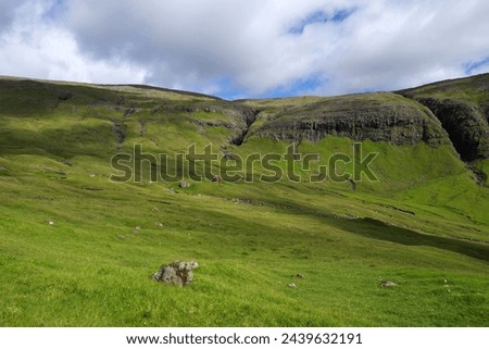 A small hut stands alone in a vast valley with a weathered, craggy cliff rising behind on the island of Vagar, Faroe Islands