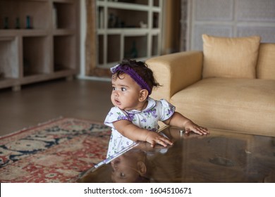 A Small Indian Girl, A Toddler In A Light Dress, Stands At A Low Coffee Table In The Interior Of The House, Holds Onto The Table And Looks Away In Surprise.