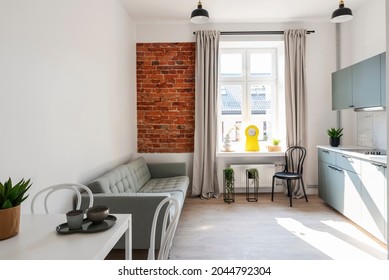 Small Interior Of Loft Apartment In Industrail Style With Window And Bricky Wall. Cozy Living Room And Kitchen With Furniture, Sofa And Table With Chairs.