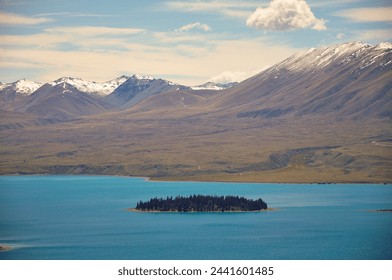 A small island in the middle of turquoise colored Lake Tekapo with snow capped mountains in the background with blue sky and clouds in New Zealand - Powered by Shutterstock