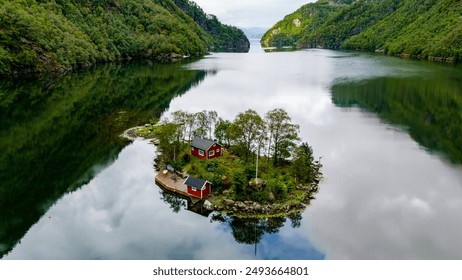 A small island with two red cabins sits nestled in a serene Norwegian fjord. Lush green trees surround the cabins, and the water reflects the surrounding landscape. Lovrafjorden, Norway - Powered by Shutterstock