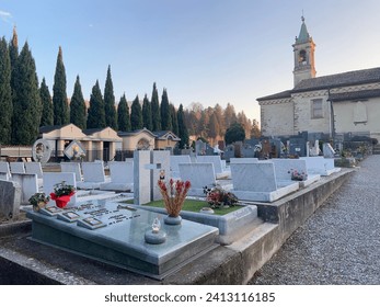 Small Italian cemetery with church in the background. Poplar trees in a row and stone wall with blue sky and clear day. Funeral monuments and the Christian religion. Crypts and village tombs.
