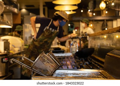 Small Japanese Restaurant With A Deep Frier In The Foreground And In The Background A Chef, And Waitresses, All Wearing Masks.