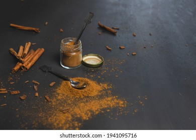 Small Jar Full Of Ground Cinnamon With A Tiny Spoon Inside And Cinnamon Sticks On The Side. On Black Background. Ceylon Cinnamon, Healthy Spice.