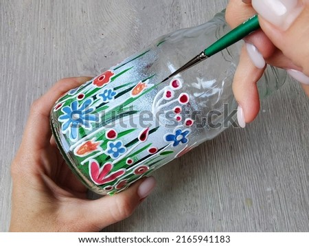 A small jar with stained glass painting. A frame from the video of the drawing master class. Artist's hands with a brush. The process of applying paint. Home hobby needlework of women housewives