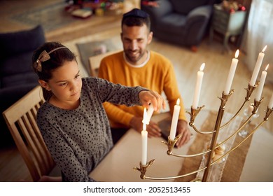 Small Jewish girl lightning up traditional candles in menorah while celebrating Hanukkah with her father at home.