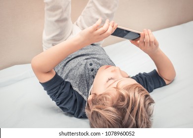 Small Kid Having Fun And Taking Selfie With Cell Phone While Lying On The Bed. 