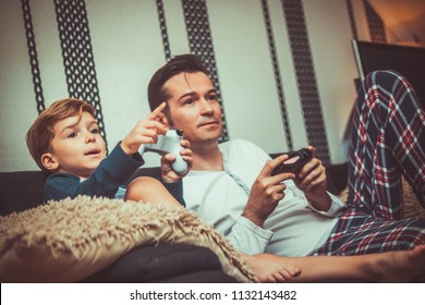 Small Kid And His Dad Playing Video Games While Relaxing On The Sofa In The Living Room.