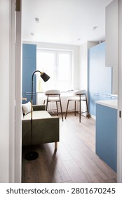 Small kitchen interior showcasing a green sofa, blue cabinets, wooden flooring, and a dining area with two stools beside a large window