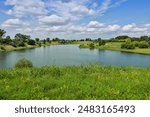 The small lake at the park under a sky with some clouds and a green field in the foreground.