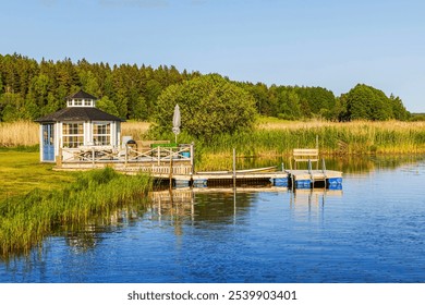 Small lakeside gazebo with wooden pier, surrounded by lush greenery under clear blue sky. Sweden. - Powered by Shutterstock