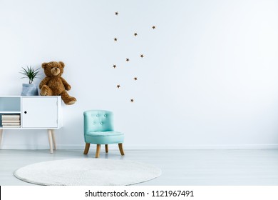 Small Light Blue Armchair For Kid Standing In White Room Interior With Stars On The Wall, White Rug And Cupboard With Books, Teddy Bear And Fresh Plant. Empty Space For Your Crib