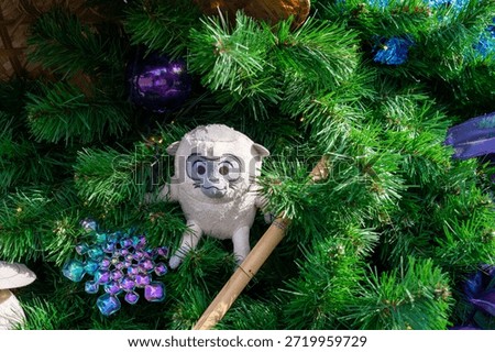 A small light colored toy monkey with long legs, big eyes, and small ears. The monkey character is hiding on a lush fir tree branch of a Christmas tree. There are purple and blue plastic decorations. The monkey has black eyes and a round face. There's a wooden stick below the figure. The colorful decorations are reflective.