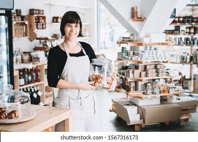 Small local business owner. Seller assistant with glass jar of pastries in interior of zero waste shop. Cheerful woman in apron stands behind counter with food products in plastic free grocery store. - Powered by Shutterstock