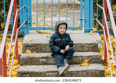 A small lonely sad child, a boy, sits on the steps in the fall near a closed kindergarten on vacation. Photography, portrait, childhood concept. - Powered by Shutterstock