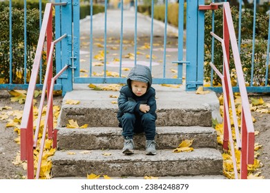 A small lonely sad child, a preschool boy, sits on the steps in the fall near a closed kindergarten on vacation. Photography, portrait, childhood concept. - Powered by Shutterstock