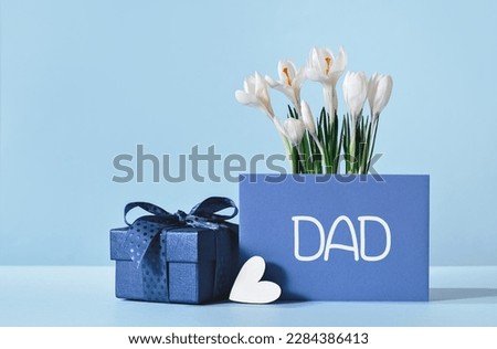 Small luxury gift box with blue bow and white heart. Dad text on gift card and white crocus flowers. Fathers day or birthday gift for him. Festive sale