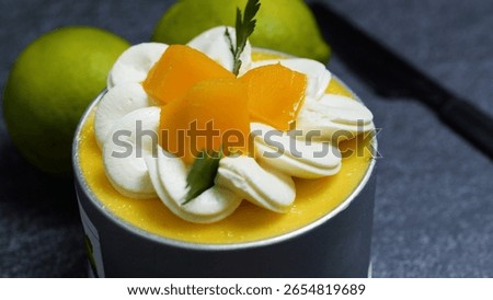 Small mango mousse cake decorated with whipped cream rosettes, mango slices, and fresh herbs, sitting on a dark gray surface with whole limes in the background