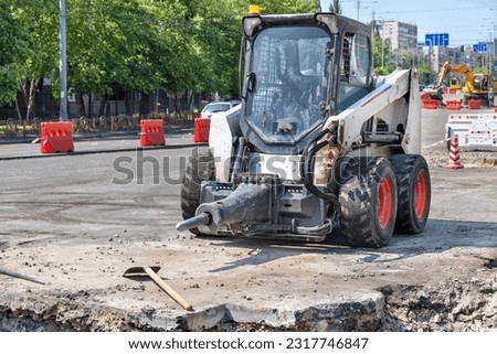 A small and manoeuvrable construction vehicle with an attached jackhammer is parked in a construction and repair area of a city roadway.