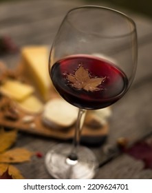 Small Maple Leaf In Glass Of Red Wine On Wooden Table With Autumn Picnic With Bread And Cheese. Romantic Dinner In Garden In Evening Outdoors. Still Life