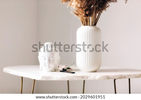 Small marble table with decorations, white vase with dry flowers, little buddha head statue and burning incense stick on stand in modern simplistic apartment environment