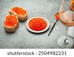 Small metal plate with red salmon caviar, two caviar toasts canape, two glasses of champagne top view on grey concrete background, festive luxury delicacy and appetizer