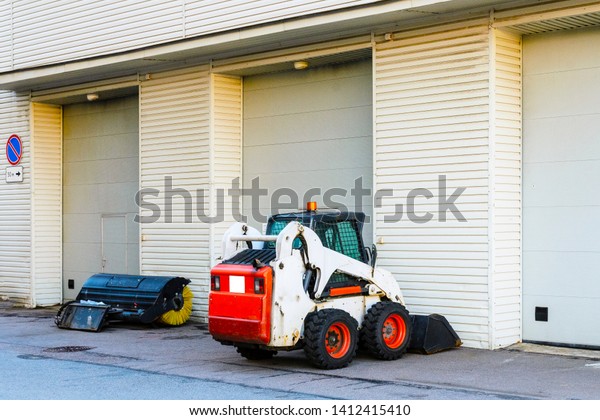 a small mini tractor with a\
blade and a bucket for street cleaning is at the gate of a large\
garage