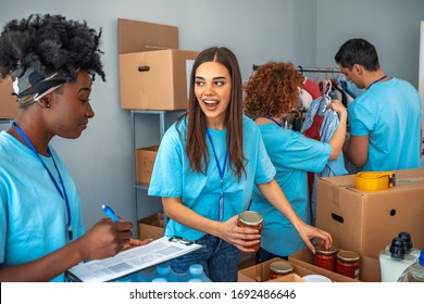 Small Mixed Race Group Of People Working In Charitable Foundation. Male And Female Teenage Food Bank Volunteers Sort Canned Food Items In Cardboard Boxes. Happy Volunteer In Community Food Bank